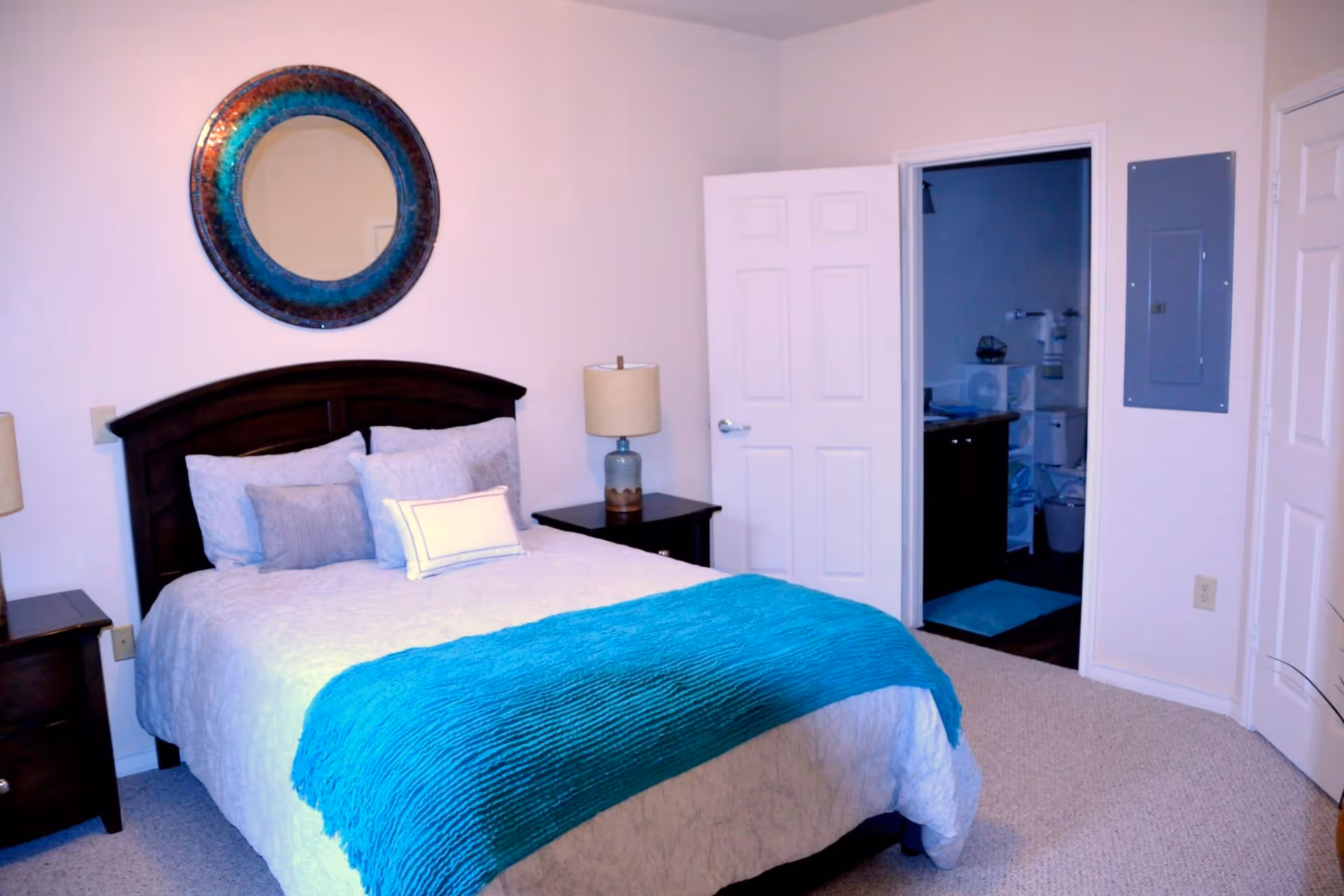 A bedroom with a neatly made bed featuring white bedding and a turquoise throw blanket. There are multiple pillows on the bed and a dark wooden headboard. On either side of the bed are matching dark wooden nightstands with lamps. A round decorative mirror with blue and brown tones hangs on the wall above the headboard. An open door reveals a glimpse of an adjoining bathroom with a blue rug and various bathroom fixtures. The room has light-colored walls and carpeted flooring.