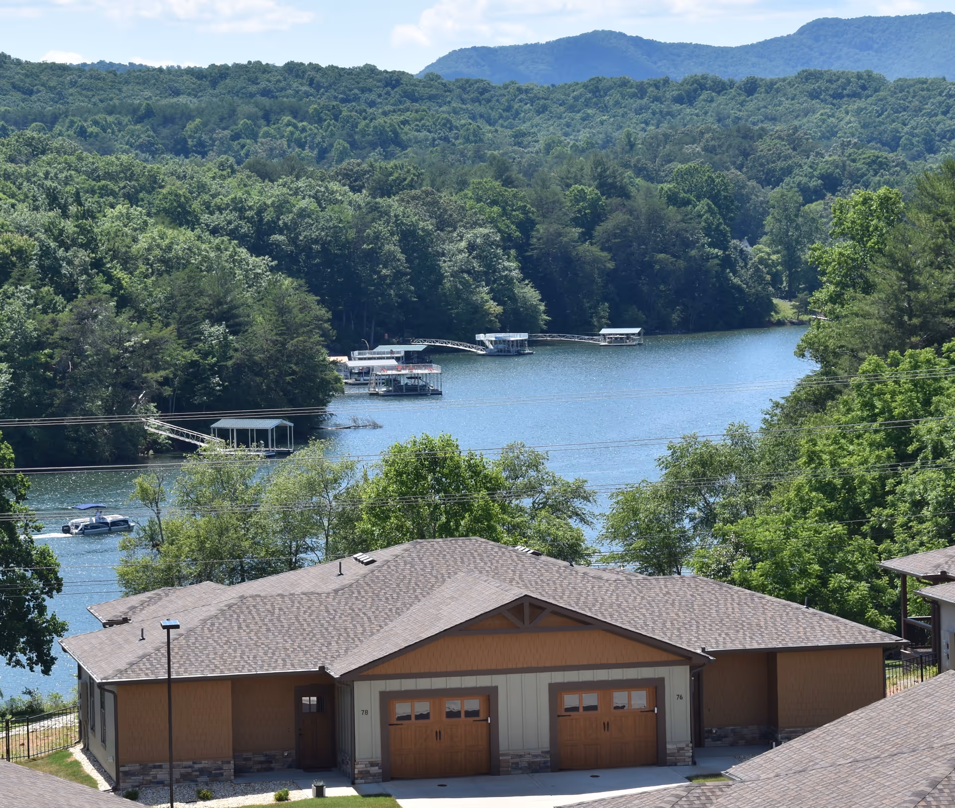 View of a residential building with two garage doors in the foreground, surrounded by trees and greenery. In the background, there is a large body of water with several boat docks and boats, and forested hills under a partly cloudy sky.