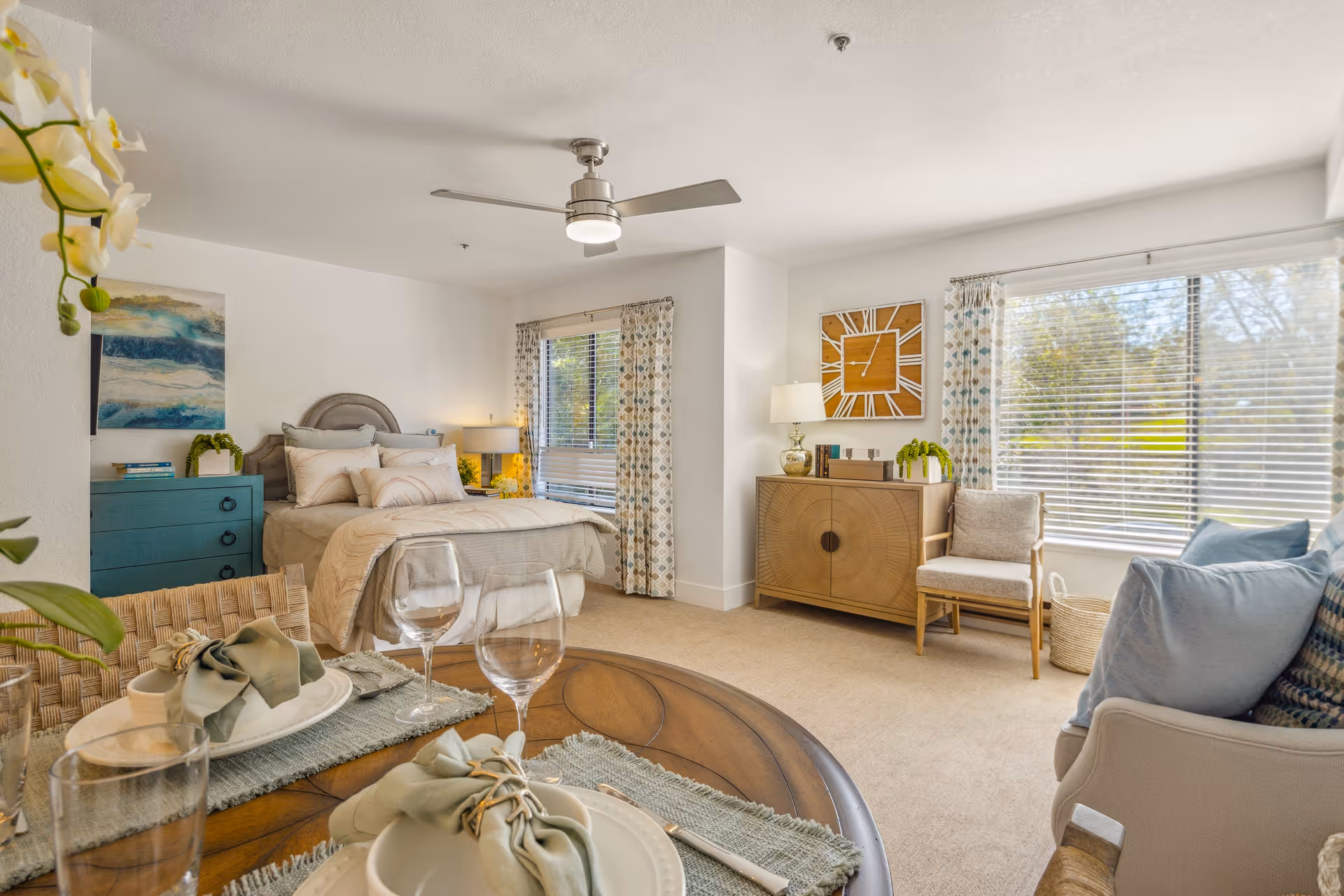 A bright and cozy studio apartment featuring a bed with beige bedding, a blue dresser, and a wooden sideboard with a lamp and decorative items. There is a dining table set with plates, glasses, and napkins in the foreground. Large windows with patterned curtains allow natural light to fill the room, and a ceiling fan is mounted above.