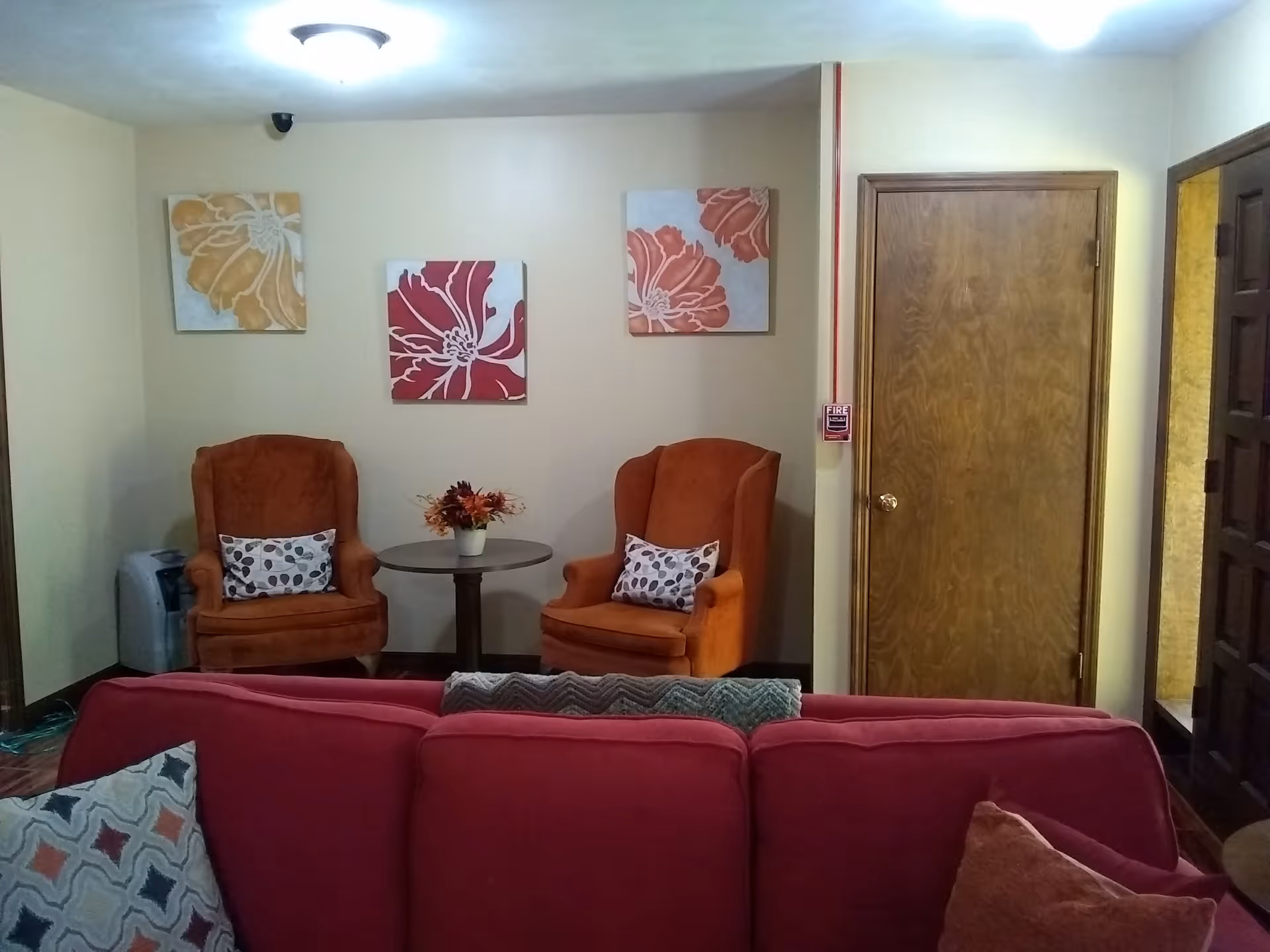 A cozy living room area with two orange armchairs, each with a decorative pillow, positioned around a small round table with a flower arrangement. Behind the chairs are three floral paintings on a beige wall. In the foreground, the back of a red sofa with patterned pillows is visible. There is a wooden door to the right and a fire alarm pull station on the wall next to it.