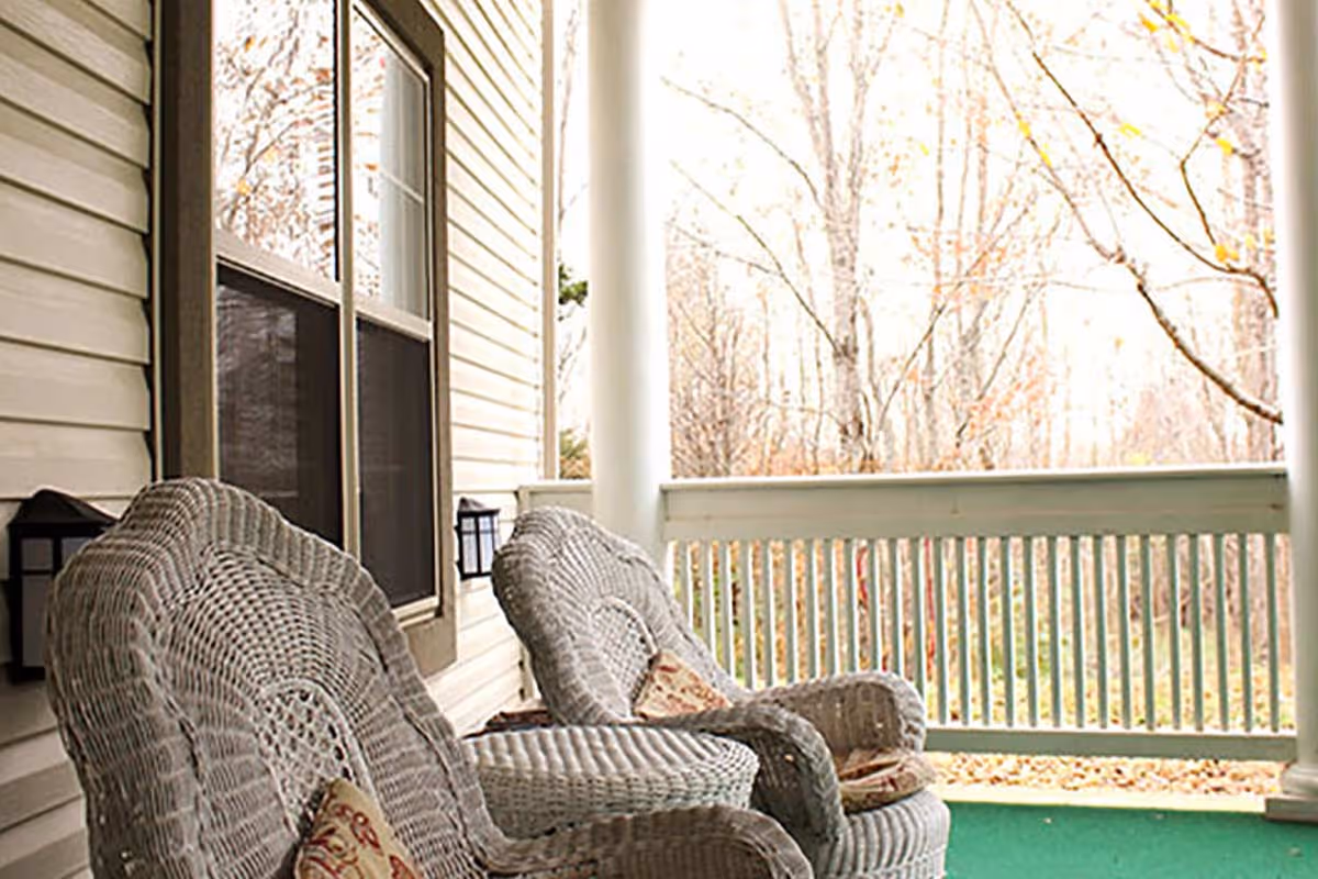 A porch area with two white wicker chairs and a matching wicker table between them. The porch has white railings and columns, with a view of leafless trees in the background.