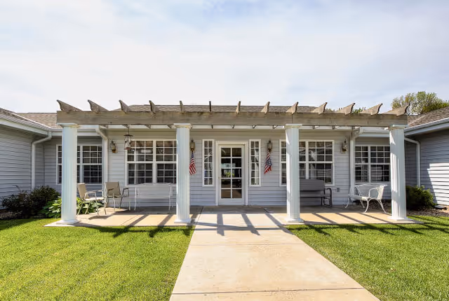 Front entrance of a single-story senior living building featuring a covered porch with white columns, seating, and American flags.