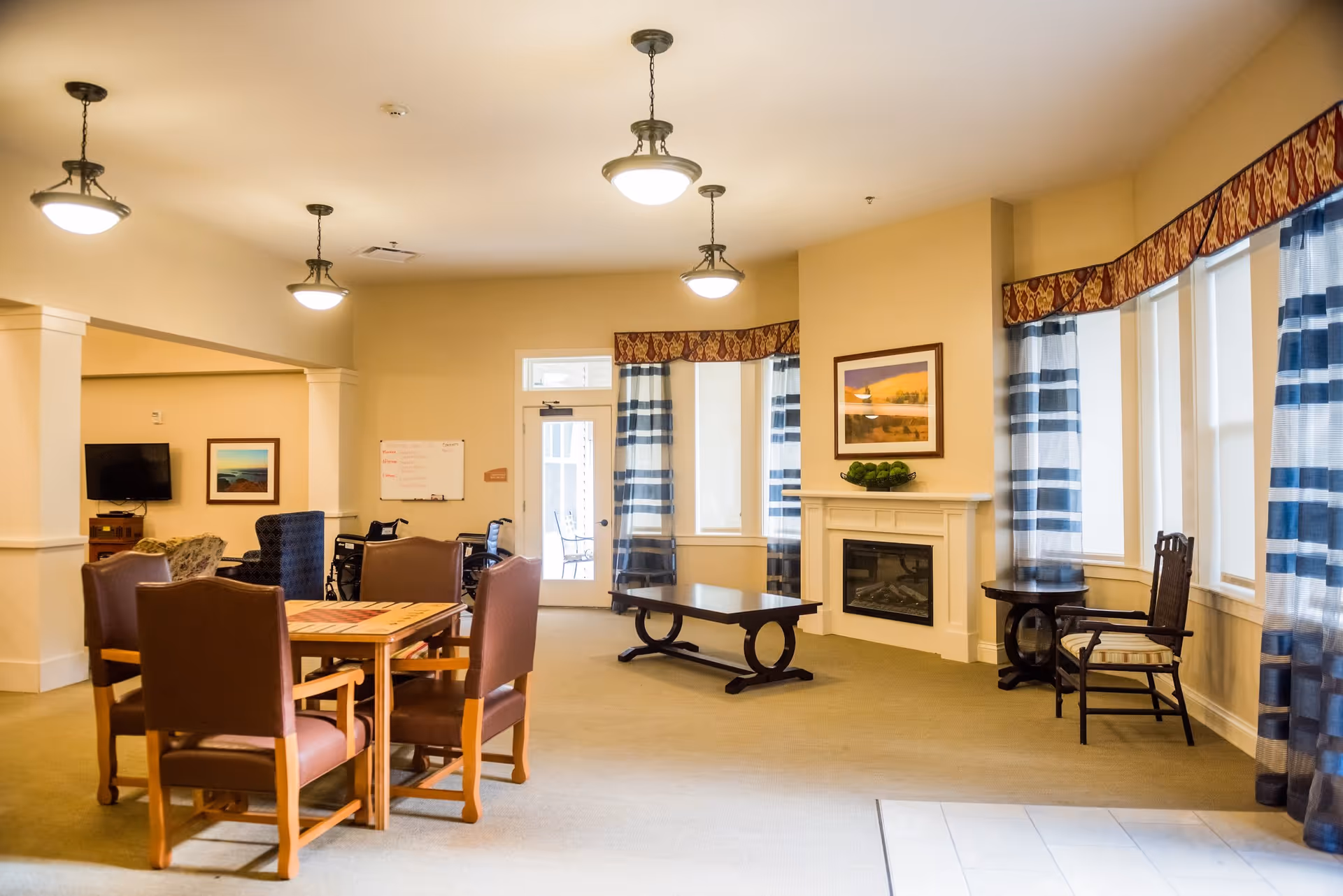 Bright communal living room with a dining table and chairs, a fireplace, TV, and large windows with striped curtains.