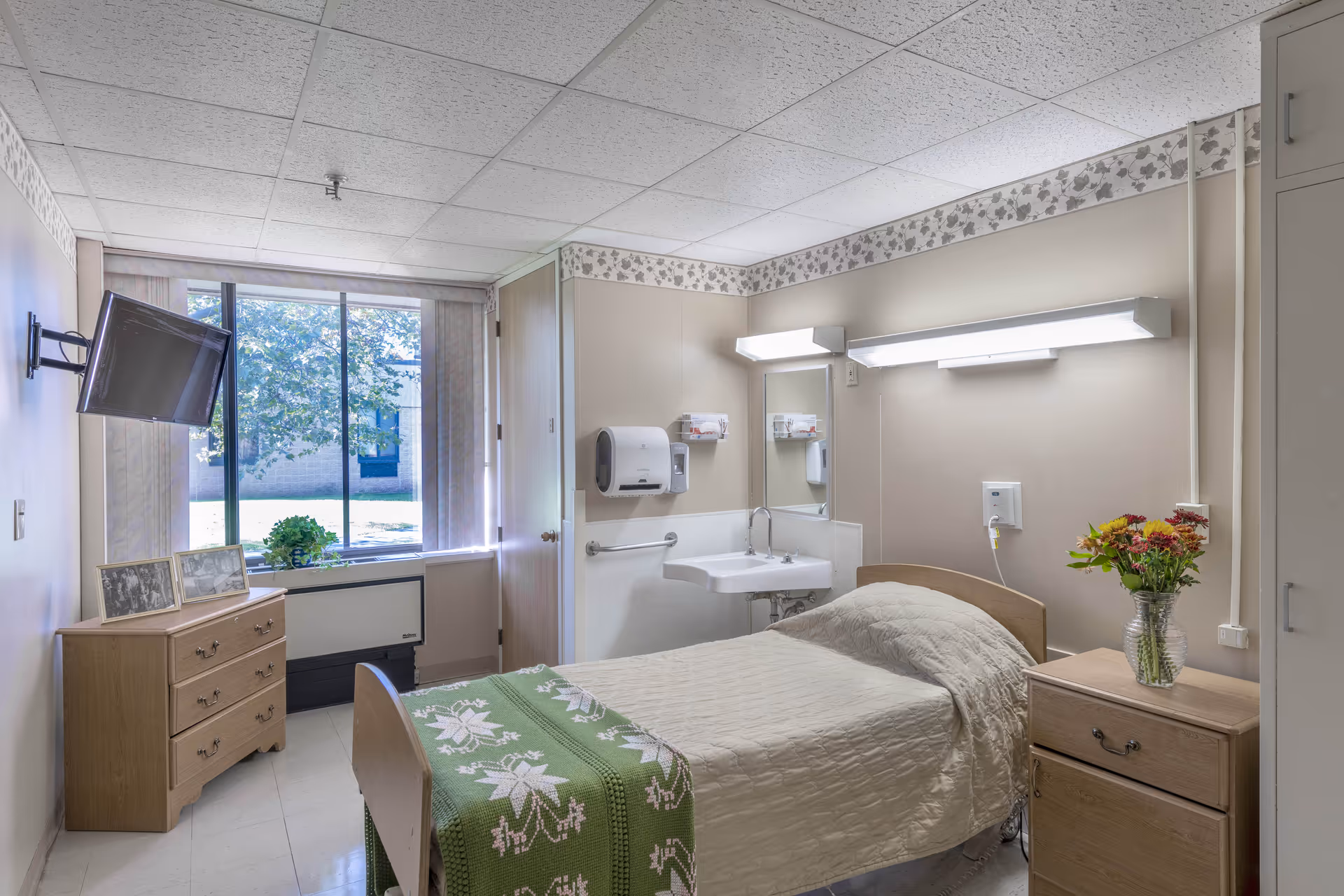 A small, neatly arranged bedroom in a senior living facility with a single bed covered in a beige quilt and a green blanket with white floral patterns. There is a wooden nightstand with a vase of colorful flowers next to the bed, and a wooden dresser with framed pictures beneath a wall-mounted TV. A large window lets in natural light, and a small sink with a mirror and paper towel dispenser is mounted on the wall. The room has beige walls with a floral border near the ceiling and a tiled floor.