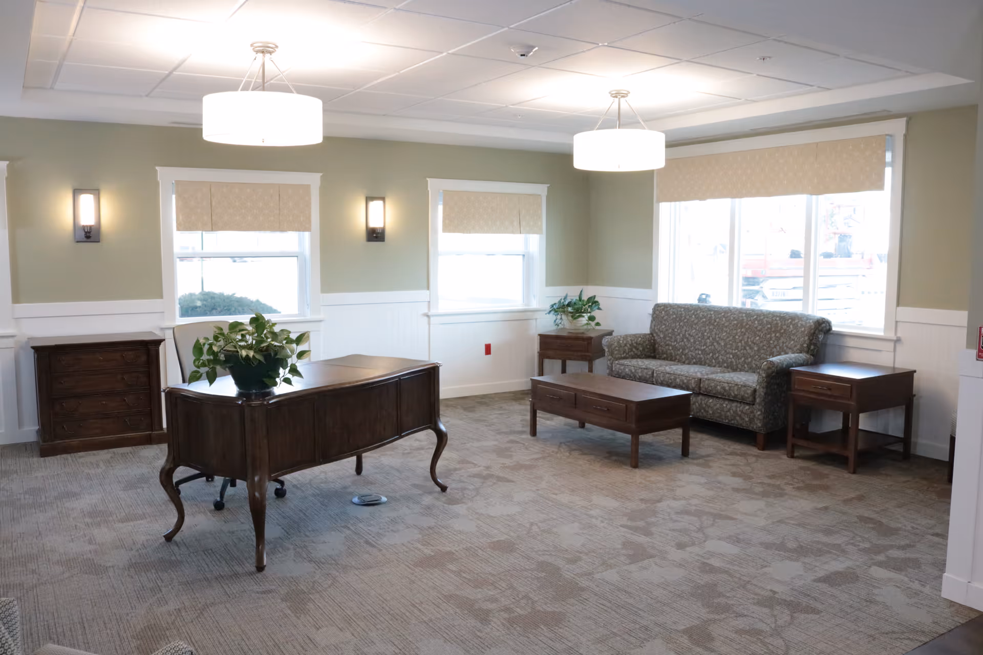 A well-lit living room area with a patterned sofa, wooden coffee table, two wooden side tables, and a wooden desk with a plant on top. The room has three windows with beige blinds, light green walls with white wainscoting, and two ceiling light fixtures.