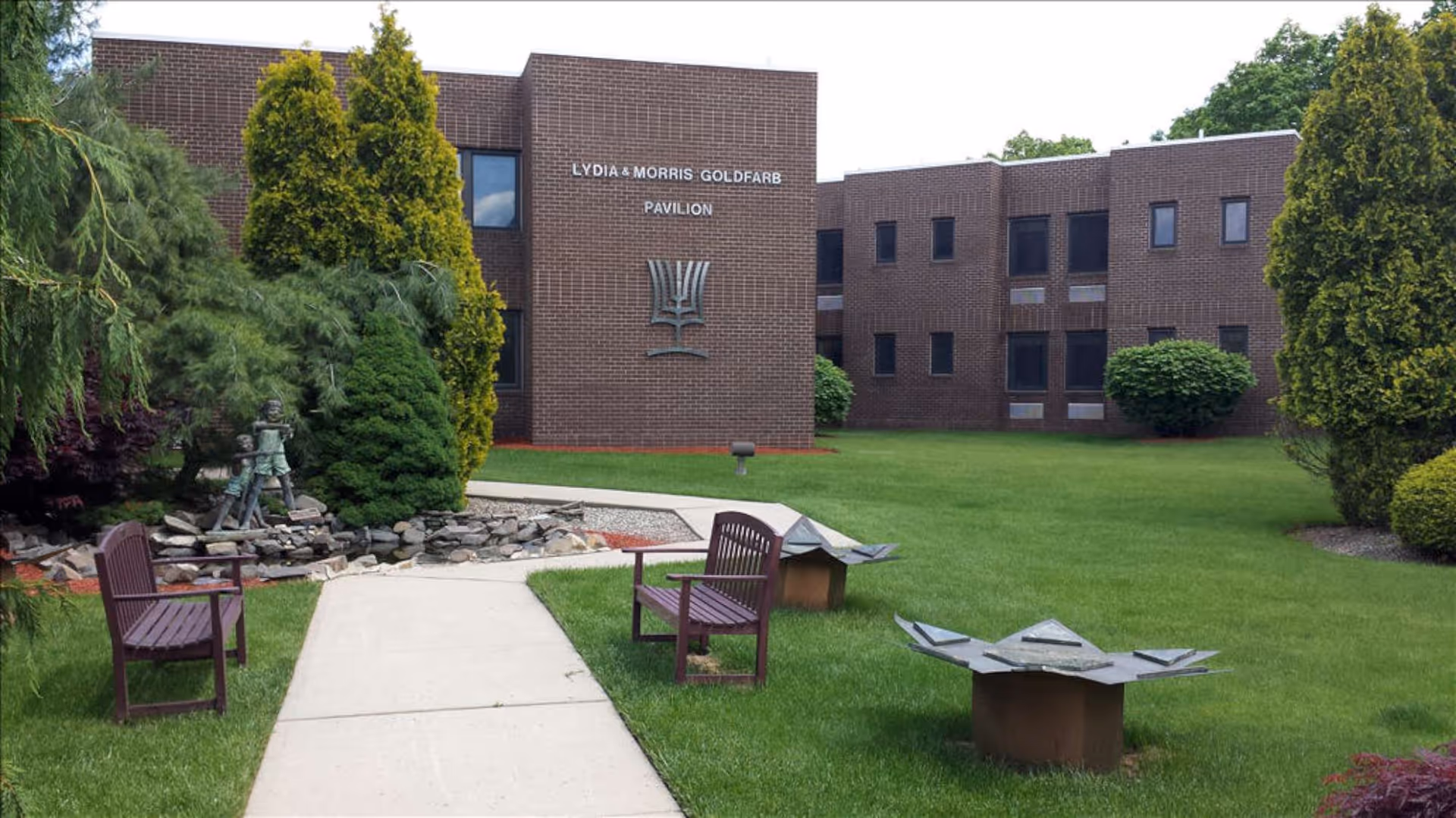 Outdoor garden area with green grass, bushes, and trees surrounding a paved walkway. Two wooden benches are placed on either side of the walkway. In the background, there is a brown brick building with the text 'LYDIA & MORRIS GOLDFARB PAVILION' and a decorative metal sculpture mounted on the wall.