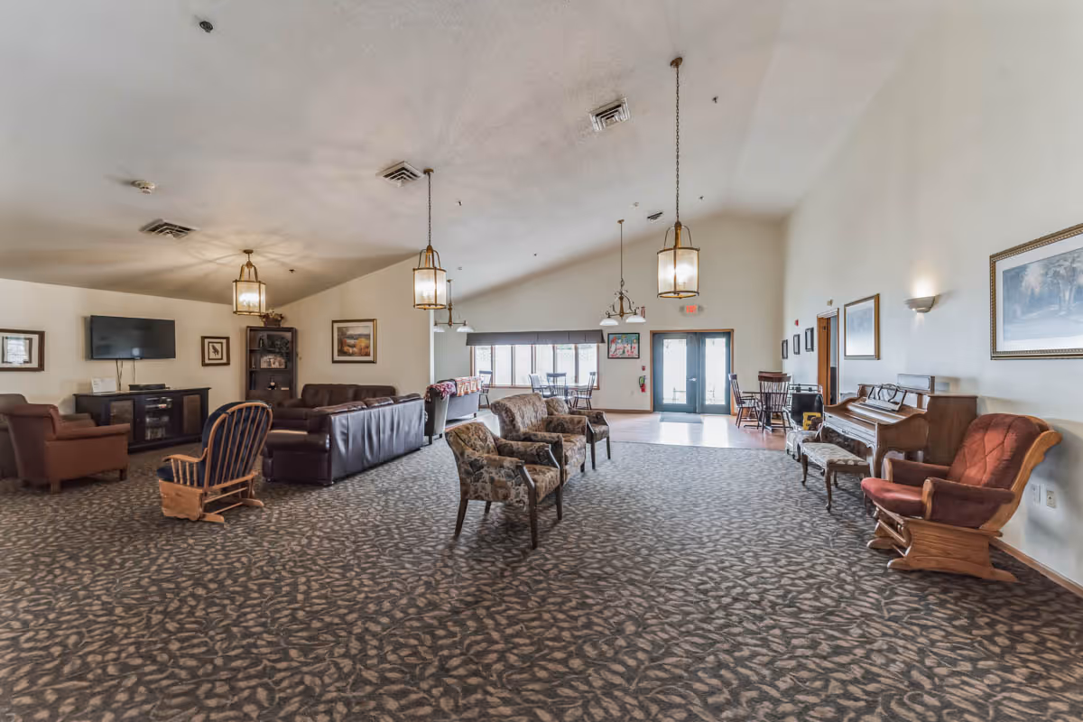 A spacious senior living facility common area with patterned carpet flooring, multiple seating options including leather sofas, armchairs, and wooden rocking chairs. The room features hanging pendant lights, a wall-mounted TV, framed artwork, a piano with a cushioned bench, and large windows letting in natural light near the entrance doors.