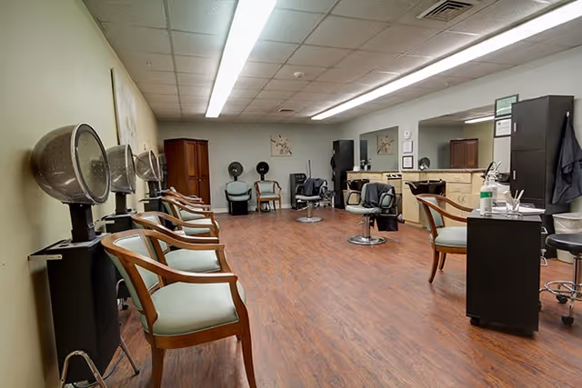 Interior view of a hair salon area with multiple hair drying stations along the left wall, wooden chairs with light green cushions, salon chairs in the center, and mirrors with countertops along the right wall. The floor is wood and the ceiling has fluorescent lighting.