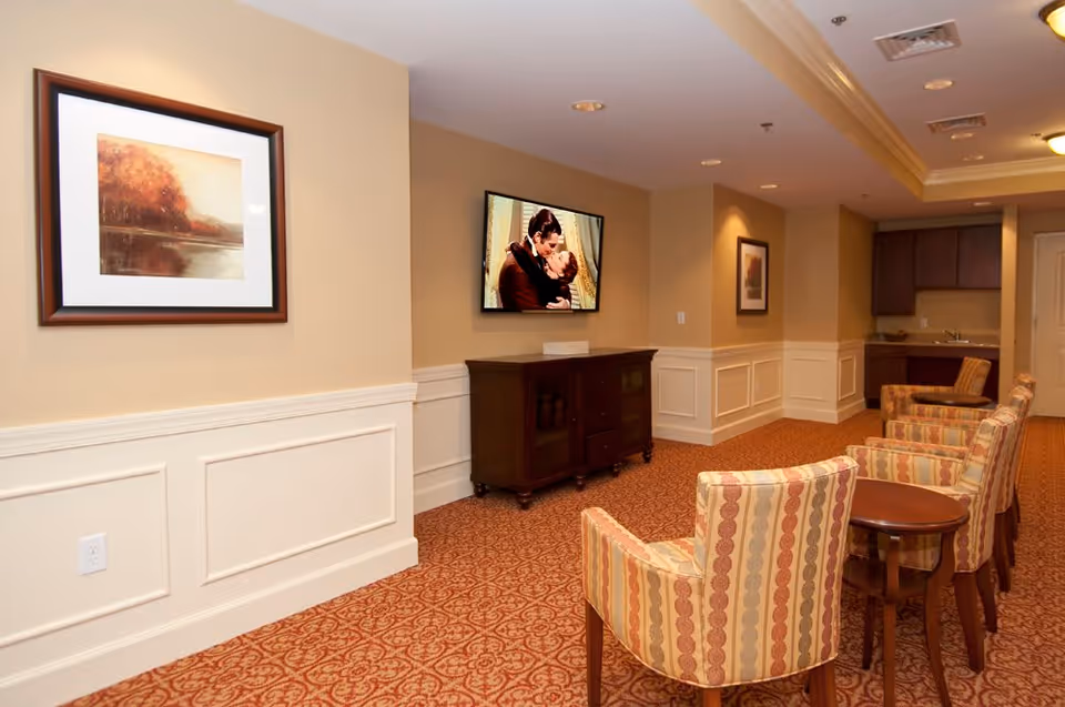 A cozy senior living common area with patterned carpet and four striped armchairs arranged in a row facing a wall-mounted TV. The TV is above a dark wooden cabinet. The walls are beige with white wainscoting and decorated with framed artwork. In the background, there is a kitchenette with dark cabinets and a sink.