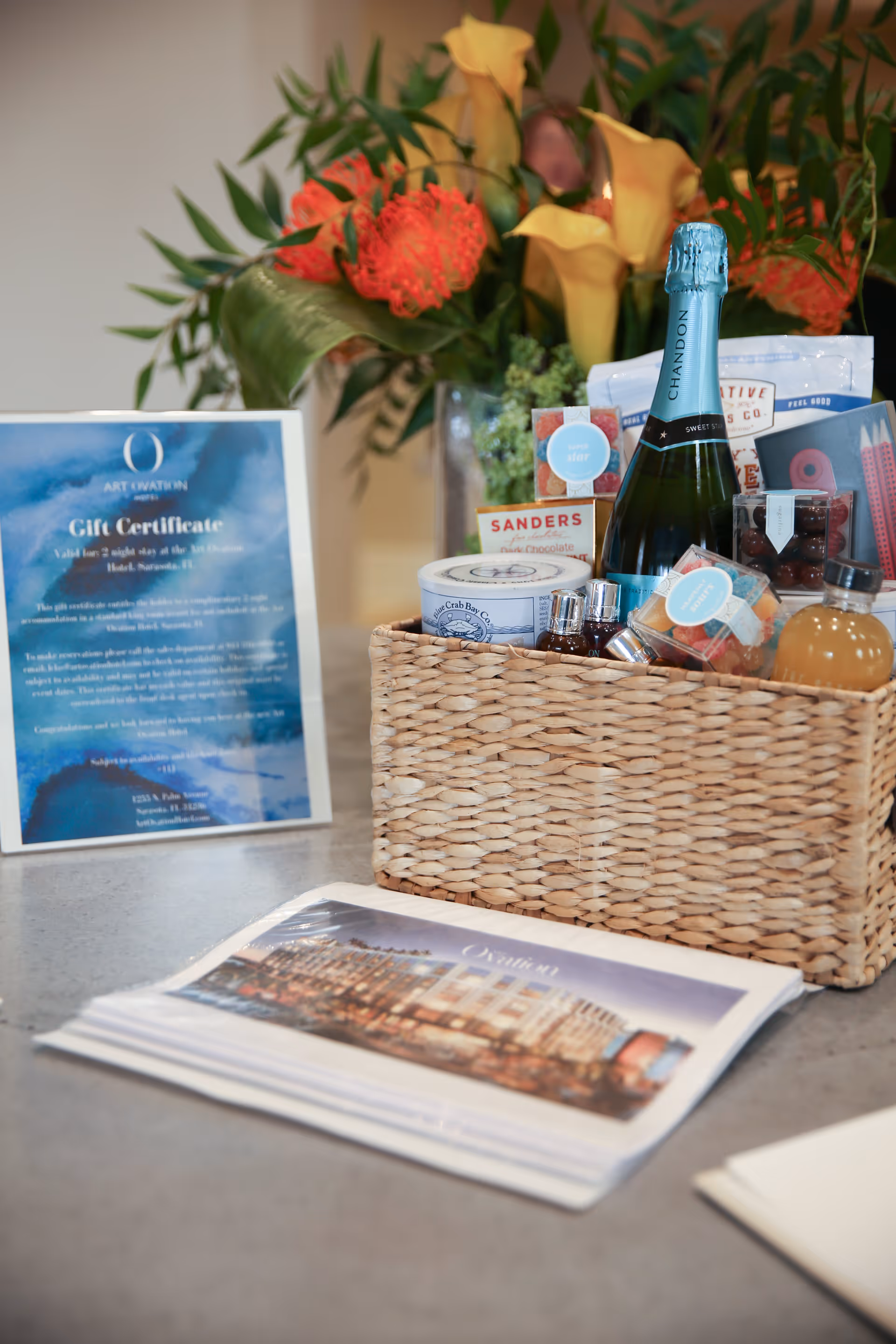 A gift basket containing a bottle of Chandon sparkling wine, assorted snacks, and small bottles of beverages sits on a countertop. Behind the basket is a framed gift certificate and a floral arrangement with orange and yellow flowers. A brochure or magazine featuring a building is also on the countertop.