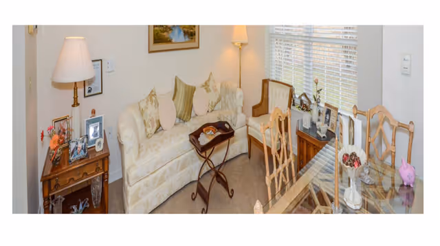 Cozy living room and dining area with a floral sofa, armchairs, side tables with lamps and framed photos, and a glass-top dining table by a window.