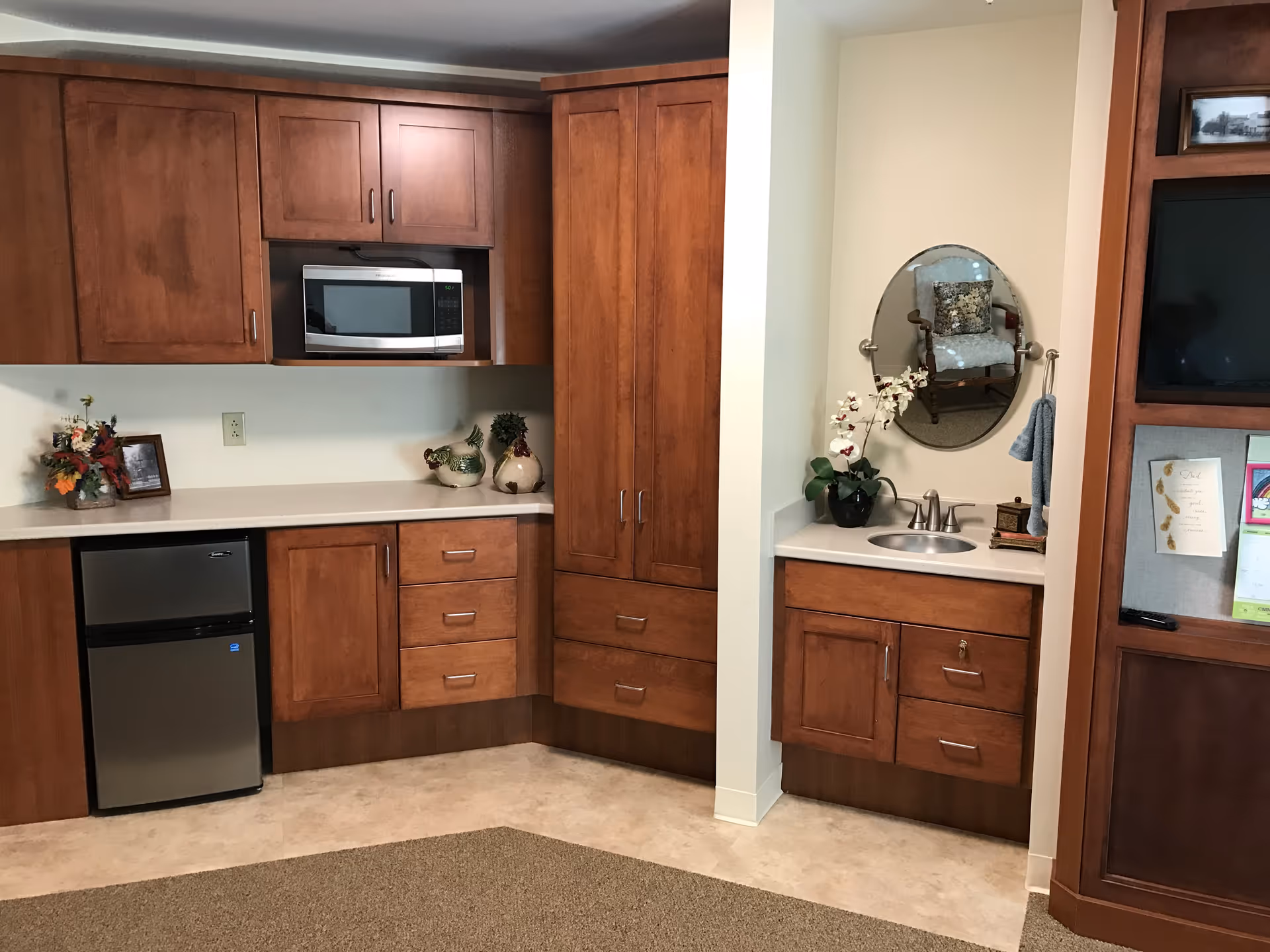 Interior view of a kitchenette area with wooden cabinets, a small stainless steel refrigerator, a microwave, and a countertop decorated with ceramic roosters and a small flower arrangement. To the right, there is a small sink with a round mirror above it and a towel hanging on the wall. A bulletin board and a TV are partially visible on the far right.