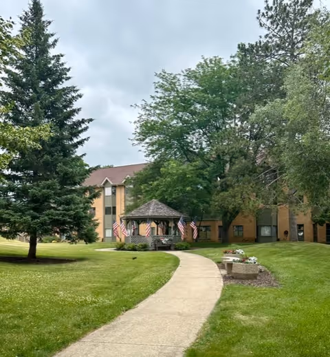 A paved walkway curves through a green lawn leading to a gazebo adorned with American flags. The gazebo is surrounded by trees and is in front of a multi-story brick building under a cloudy sky.