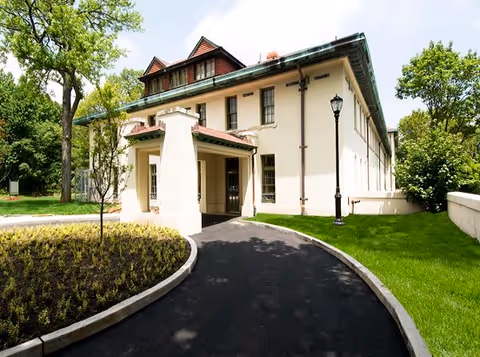 Exterior view of a two-story building with a covered entrance, surrounded by green grass, trees, and a curved paved driveway under a partly cloudy sky.