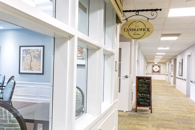 Interior hallway of a senior living facility with a sign hanging that reads 'Candlewick bistro' and a chalkboard listing lunch specials. To the left, a dining area with tables, chairs, and framed artwork on the wall is visible through large windows.