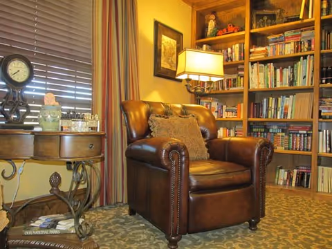 Cozy corner of a living room featuring a brown leather armchair with a decorative pillow, a wooden side table with a clock and decorative items, a floor lamp, and a bookshelf filled with books and small decorative objects.