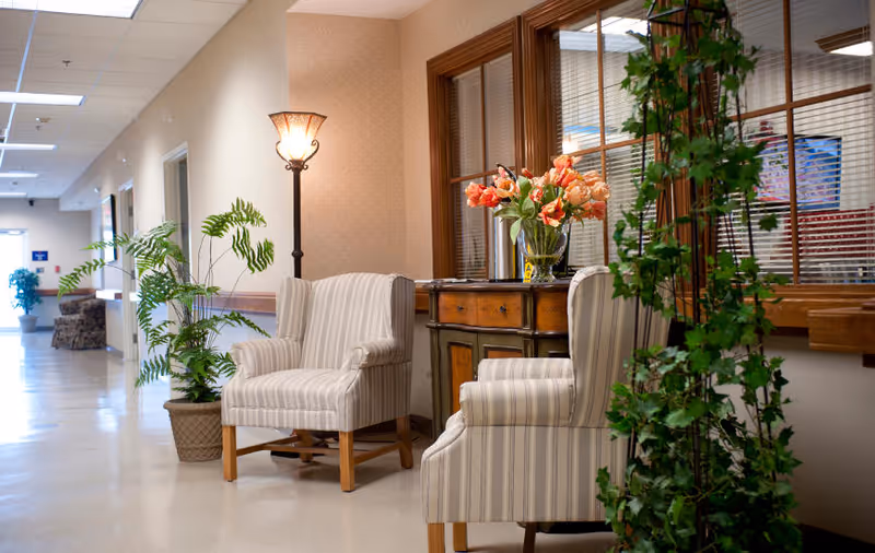 A well-lit hallway in a senior living facility with two striped armchairs, a wooden side table with a vase of orange flowers, a tall floor lamp, and several potted plants. The hallway has beige walls, a tiled floor, and large windows with wooden frames on the right side.