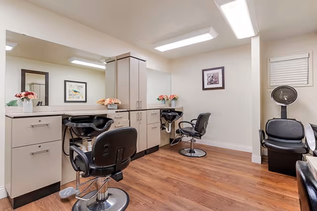 Interior view of a hair salon area in a senior living facility with two black salon chairs in front of sinks and mirrors, beige cabinets, wooden flooring, and a hair dryer chair in the corner. The walls are light-colored with framed artwork and a small window with blinds.