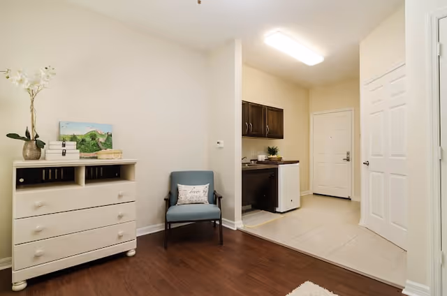 Interior view of a room in American House Bonita Springs Independent Living featuring a white dresser with decorative items including a vase with flowers and a painting, a blue armchair with a pillow, and a small kitchenette area with dark cabinets, a mini fridge, and a countertop near the entrance door.