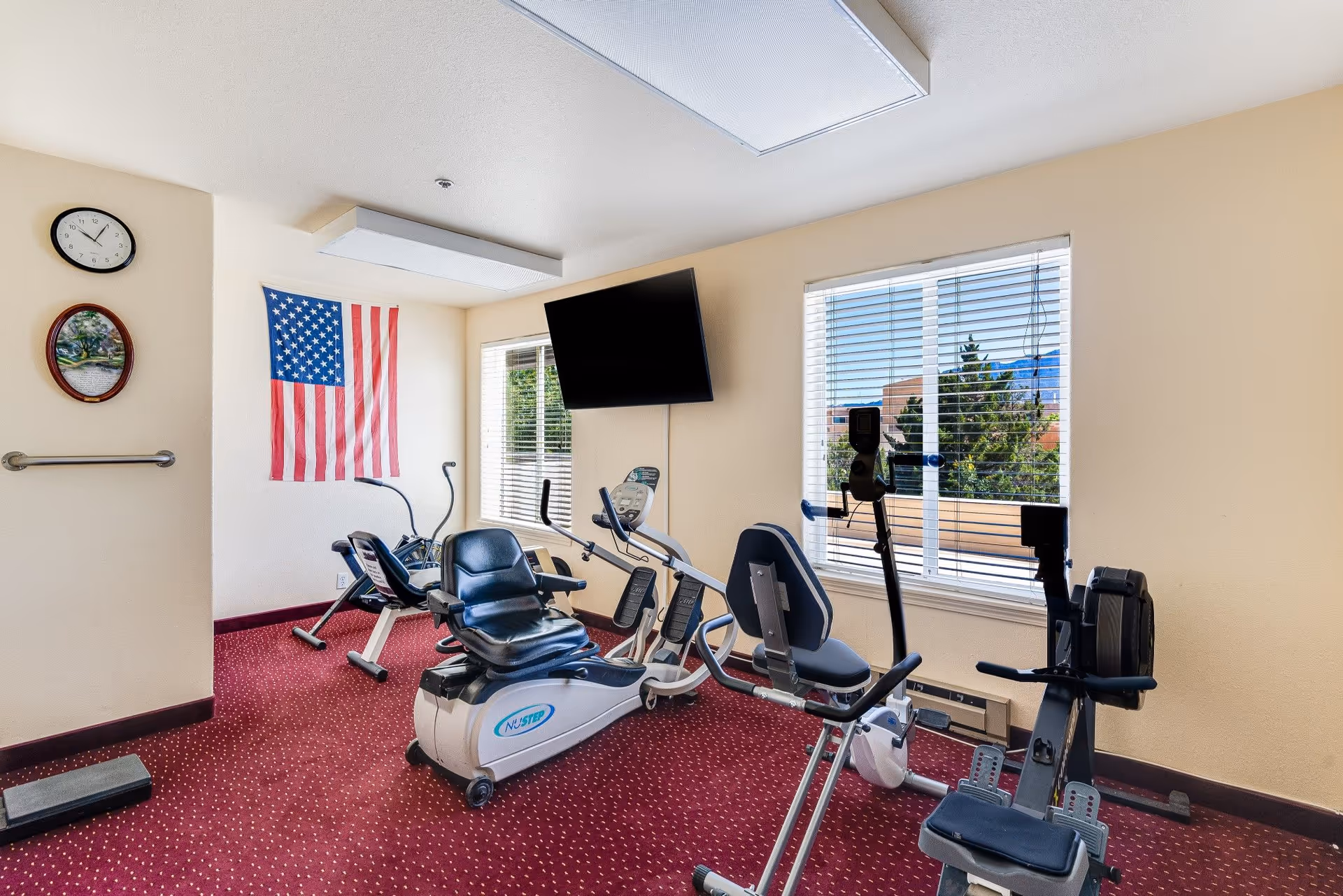 A small exercise room with red carpet and beige walls featuring three pieces of exercise equipment including a recumbent bike and a rowing machine. There is an American flag hanging on the wall, a clock, a framed picture, and a flat-screen TV mounted on the wall. Two windows with blinds let in natural light and show trees and buildings outside.