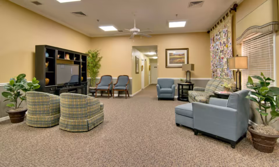 A cozy senior living facility common area with patterned carpet, beige walls, and ceiling lights. The room features a black entertainment center with a TV, two patterned swivel chairs, two blue armchairs, a striped sofa, side tables with lamps, potted plants, and a colorful quilt hanging on the wall. A hallway is visible in the background.