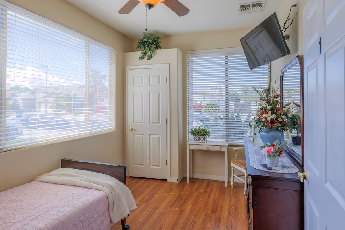 A small, bright bedroom with a single bed covered in a pink quilt and a beige blanket. The room has two large windows with white blinds, a ceiling fan, a white door, a small white desk with a plant on it, and a dark wooden dresser with a mirror and flower arrangements. A flat-screen TV is mounted on the wall above the dresser.