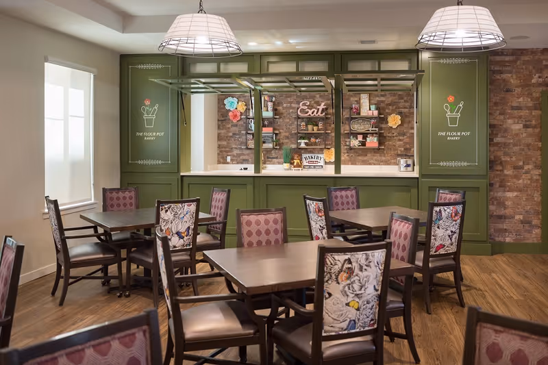 A cozy dining area with several wooden tables and chairs featuring patterned upholstery. The back wall has green paneling with decorative signs that read 'THE FLOUR POT BAKERY' and a brick accent wall with shelves holding various decorative items and a sign that says 'Eat'. Two pendant lights hang from the ceiling above the tables.