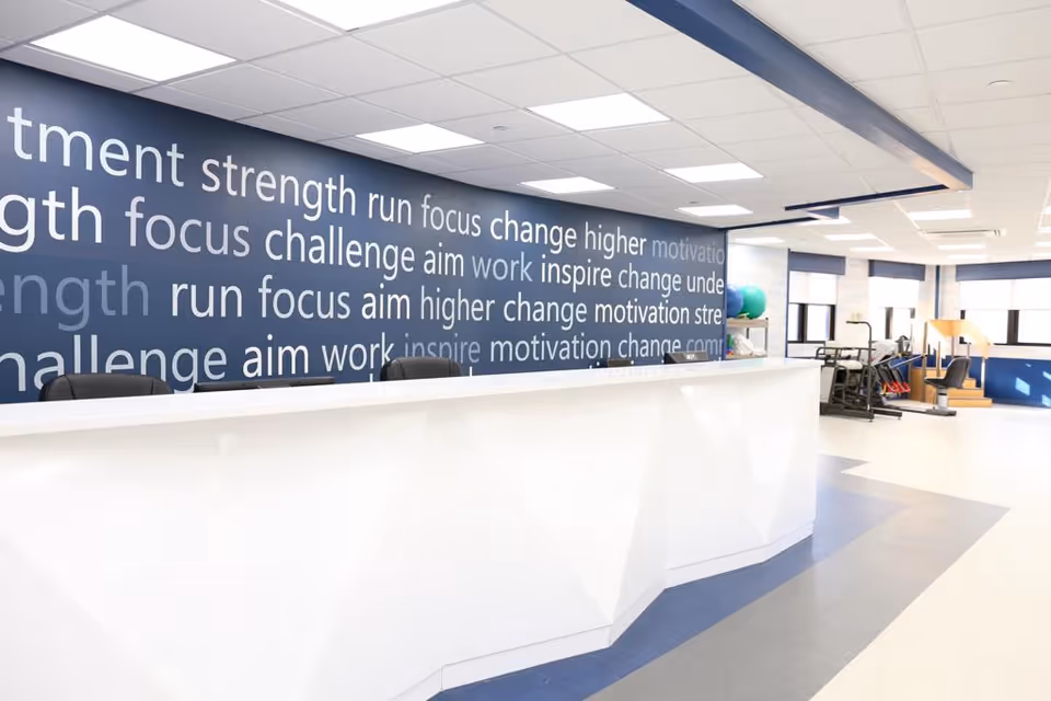 Bright interior reception area with a white desk, a blue feature wall covered in motivational words, and therapy equipment visible in the background.