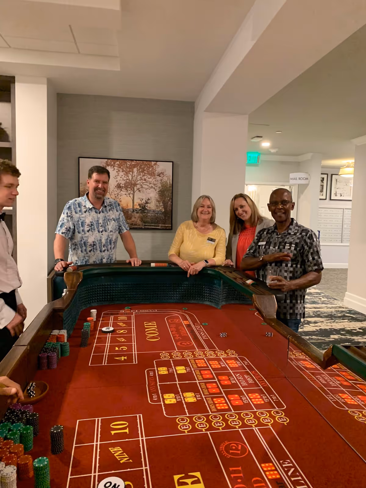 A group of five adults standing around a craps table indoors, smiling and engaging in a game. The room has neutral-colored walls with a framed picture of a tree on the wall behind them. There is a sign indicating a mail room in the background.