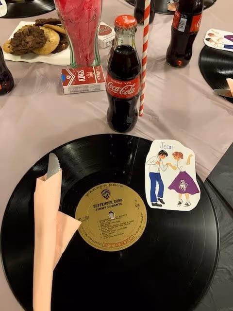A table setting featuring a black vinyl record used as a placemat, a pink cloth napkin folded with a silver utensil inside, a small illustrated place card with a dancing couple labeled 'Jean', a glass bottle of Coca-Cola with a red and white striped straw, and a plate of assorted cookies in the background.