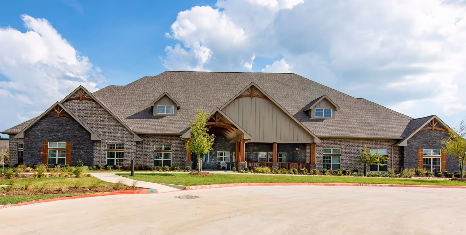 Exterior front view of a single-story assisted living and memory care facility building with a large sloped roof, multiple windows with wooden shutters, a covered entrance supported by wooden beams, and landscaped greenery in front under a partly cloudy sky.