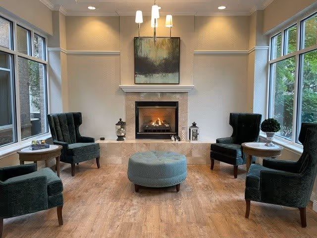 A cozy senior living common area with four dark green armchairs arranged around a round blue ottoman. The room features large windows on two sides letting in natural light, a fireplace with a fire burning, and a painting above the fireplace. There are small side tables next to the chairs with decorative items and books.