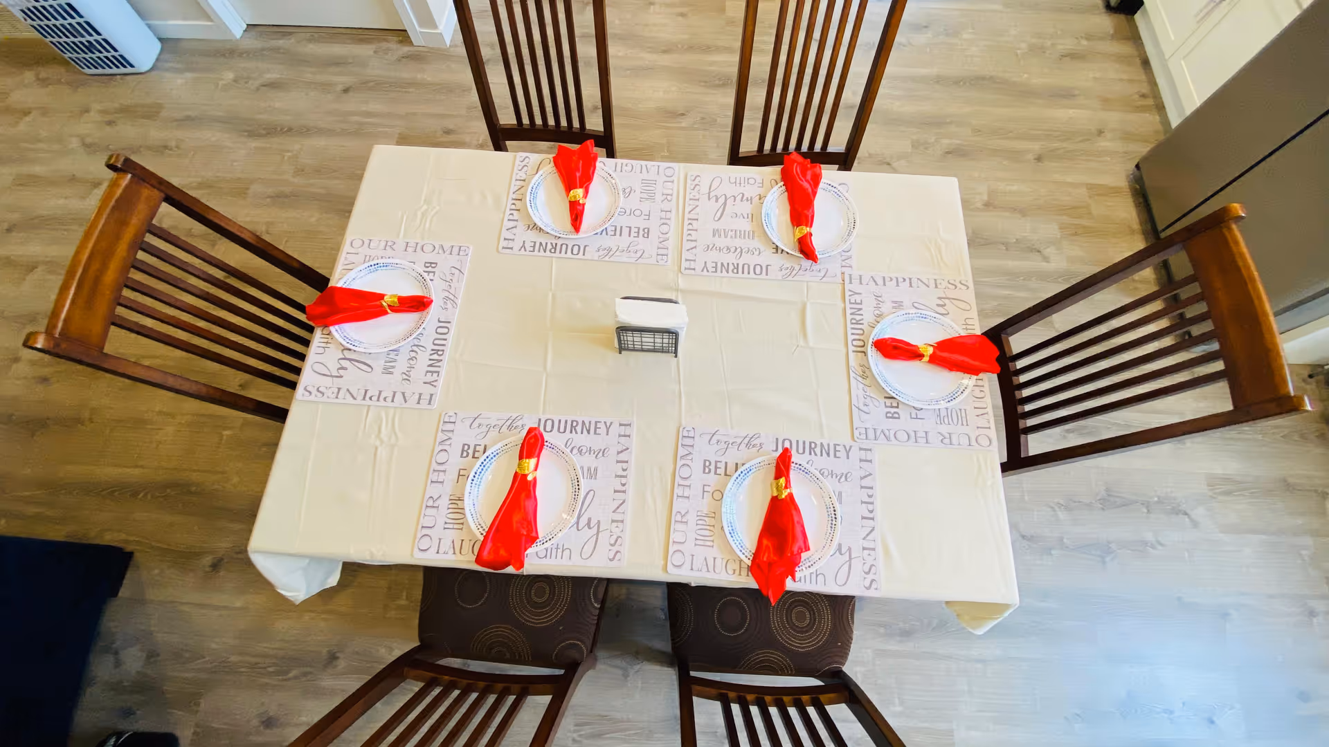 A dining table set for five people with white plates and red napkins folded and held with gold napkin rings. The table is covered with a beige tablecloth and placemats with words like 'Journey', 'Happiness', and 'Our Home'. Five wooden chairs surround the table, and the floor is light wood. A refrigerator is visible in the background.