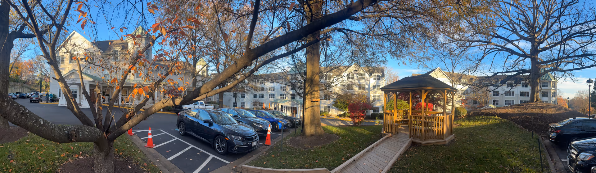 Panoramic view of the exterior of Sunrise at Bluemont Park senior living facility showing a parking lot with several cars, a wooden gazebo on a grassy area, and large trees with autumn leaves. The multi-story white building with green roofs is visible in the background under a clear blue sky.