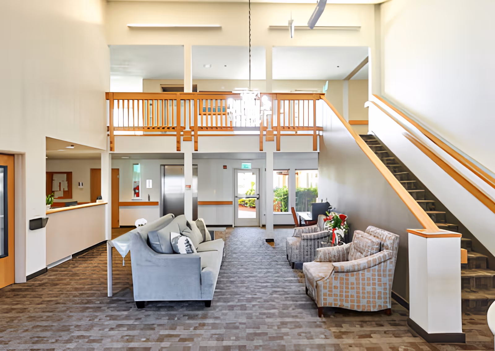Bright two-story senior living facility lobby with sofas and armchairs, a staircase and balcony overlooking the entrance and elevator.