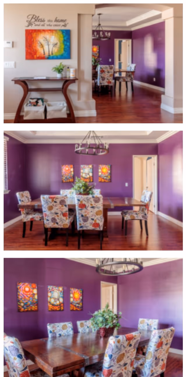 Dining room with purple walls, a wooden table surrounded by floral-upholstered chairs, a chandelier, and colorful wall art.