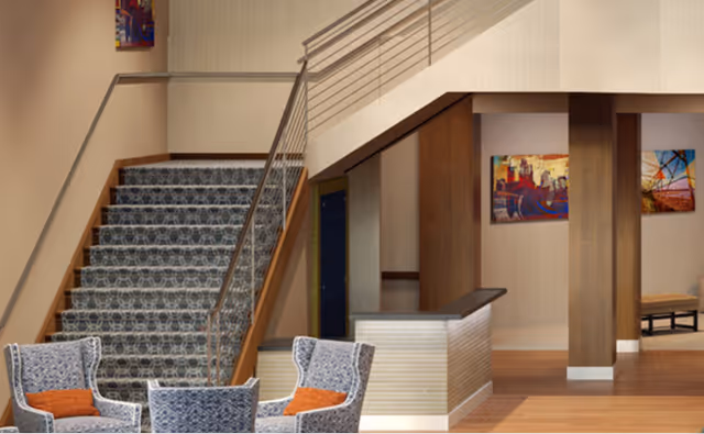 Lobby interior with a carpeted staircase, a small reception desk and upholstered chairs with orange pillows.