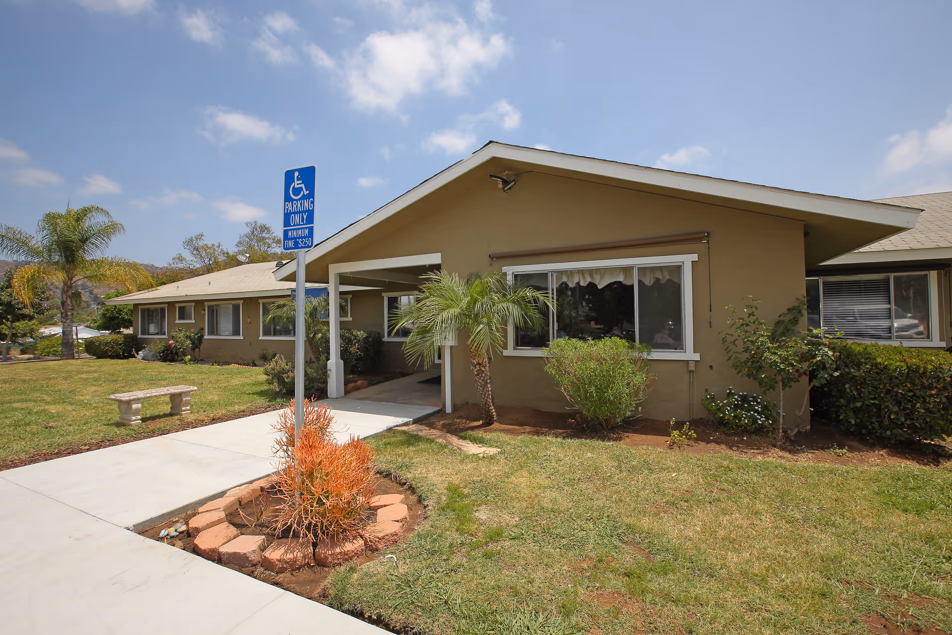 Exterior view of a single-story building with beige walls and a sloped roof, surrounded by a well-maintained lawn with small palm trees and shrubs. A concrete walkway leads to the entrance, which is covered by a small porch. A blue handicap parking sign is visible near the walkway. The sky is partly cloudy.