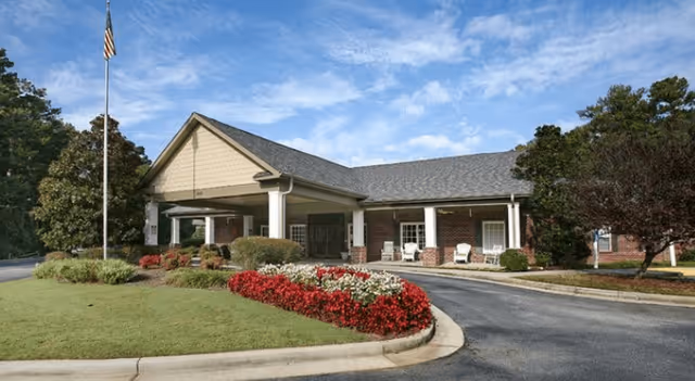 Exterior view of The Carriages of Mt. Zion facility showing a single-story brick building with a covered entrance, a flagpole with an American flag, landscaped bushes, and flower beds under a partly cloudy sky.