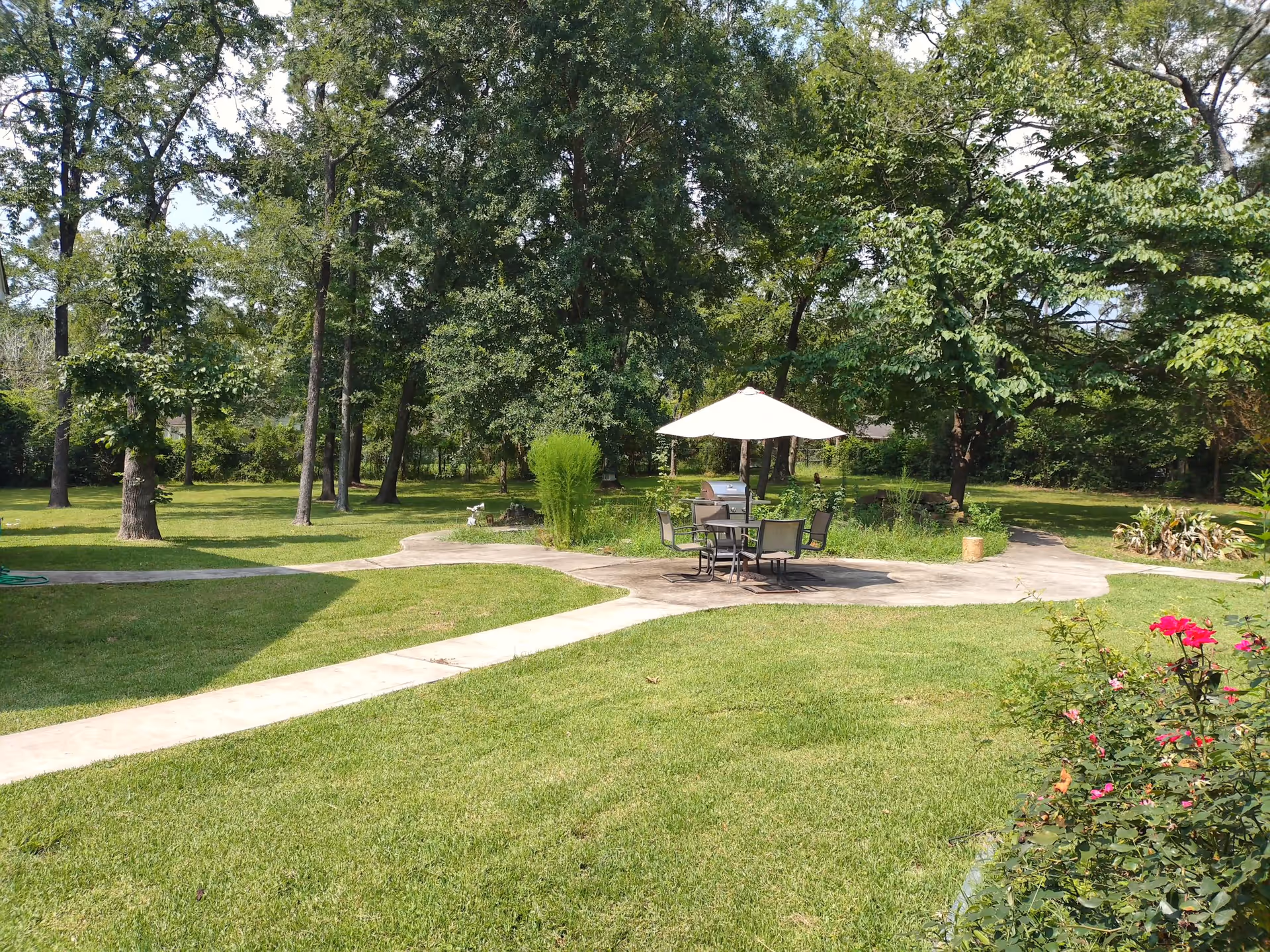 Outdoor garden area with a patio table and umbrella on a paved circle surrounded by grass, trees, and flowerbeds.