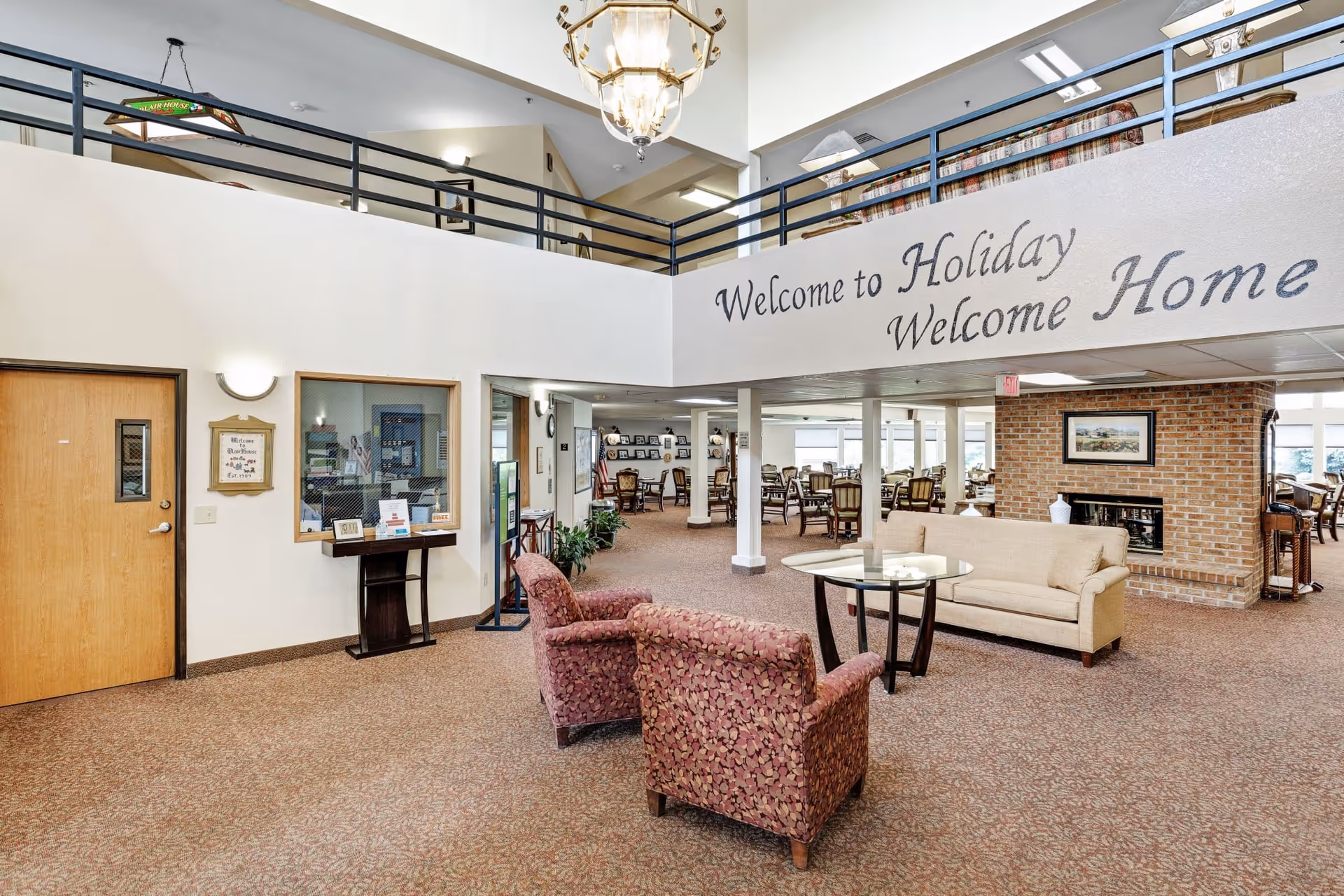 Interior view of a senior living facility lobby area with a beige sofa, two patterned armchairs, a glass coffee table, and a brick fireplace. Above, a balcony railing overlooks the space, and a sign on the wall reads 'Welcome to Holiday Welcome Home'. The area has carpeted floors, light-colored walls, and a chandelier hanging from the ceiling.