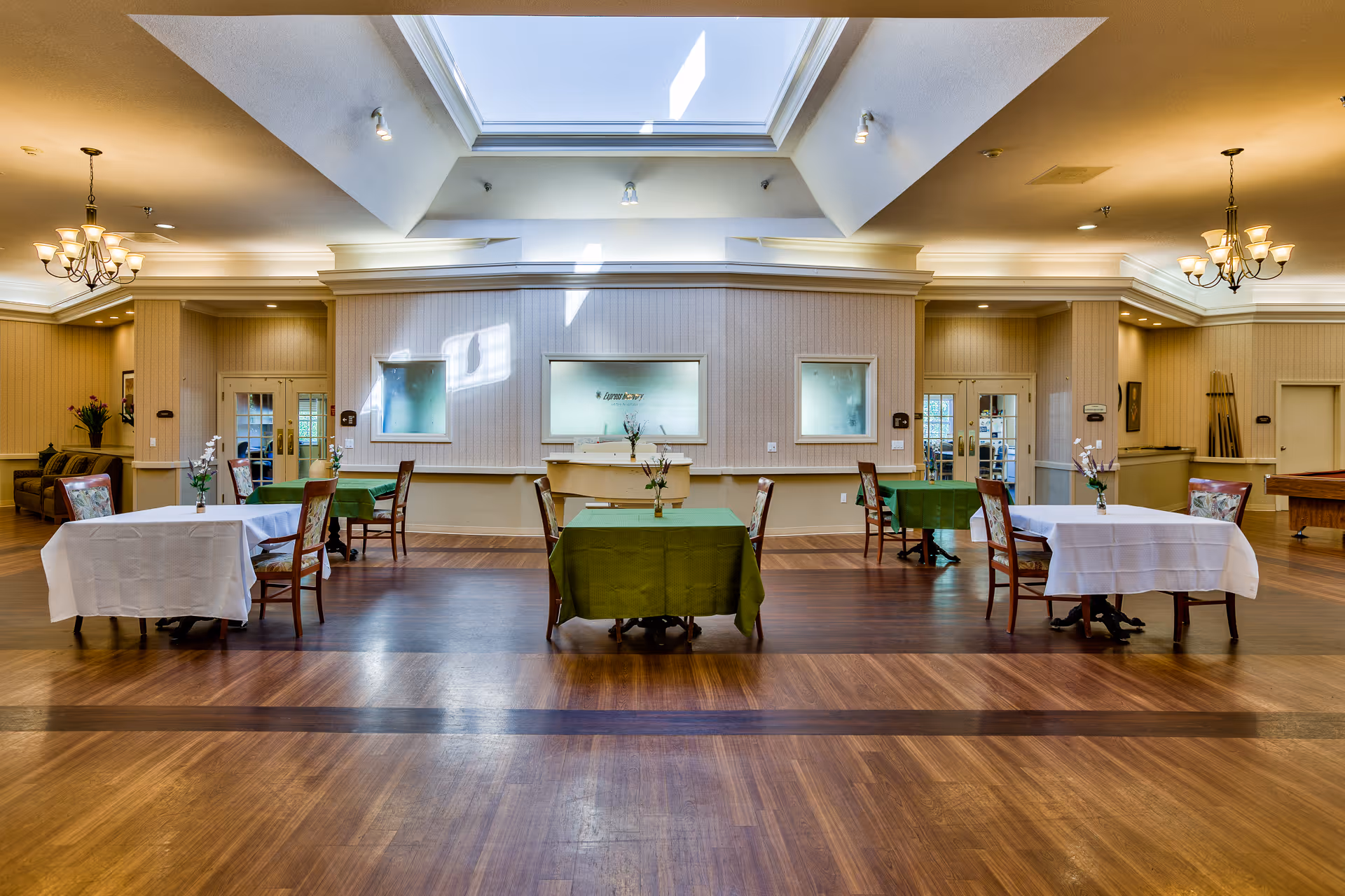 Spacious communal dining/activity room with several small tables and chairs under a large skylight.