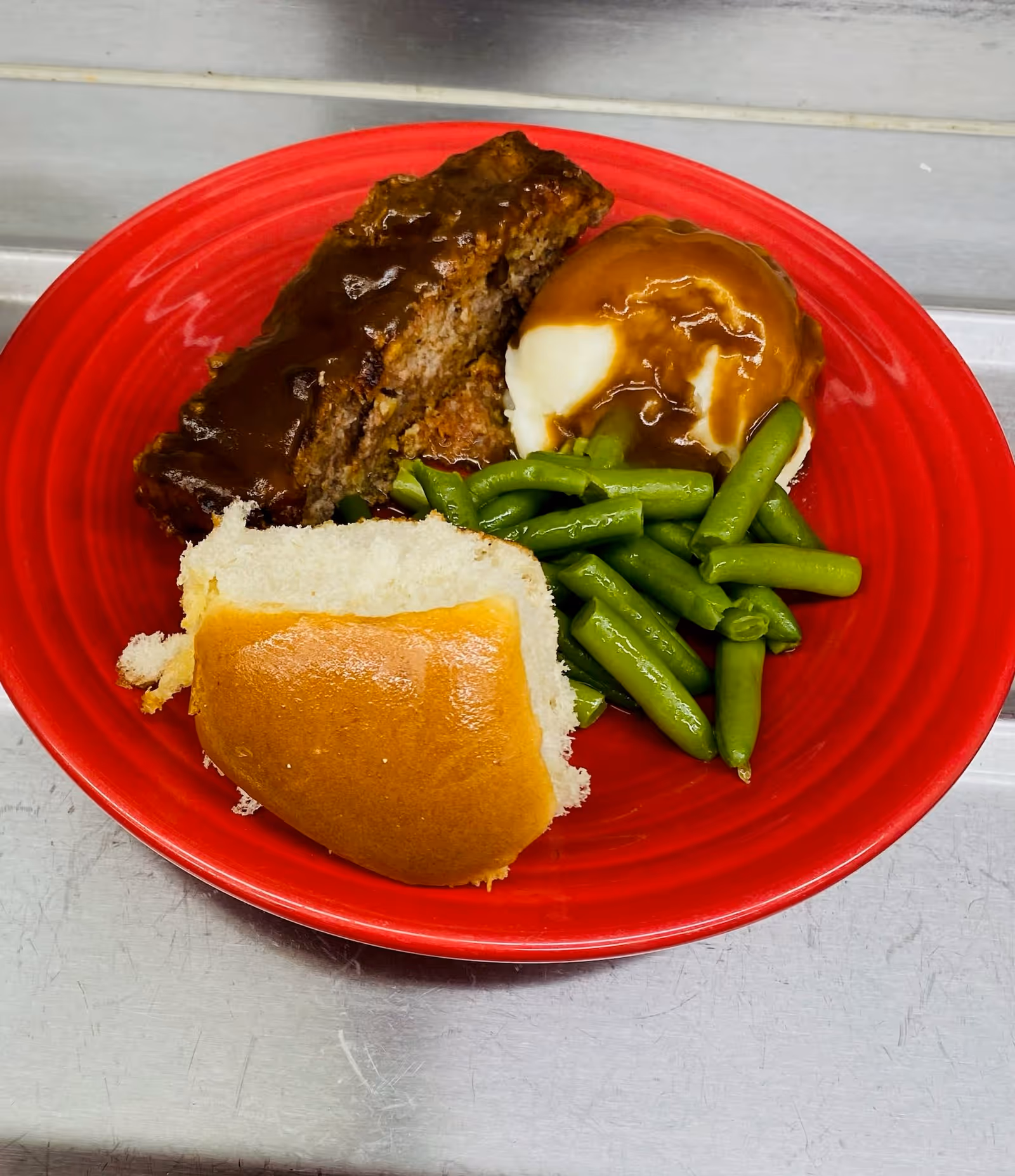 A red plate with a serving of meatloaf topped with brown gravy, mashed potatoes with brown gravy, steamed green beans, and a bread roll.