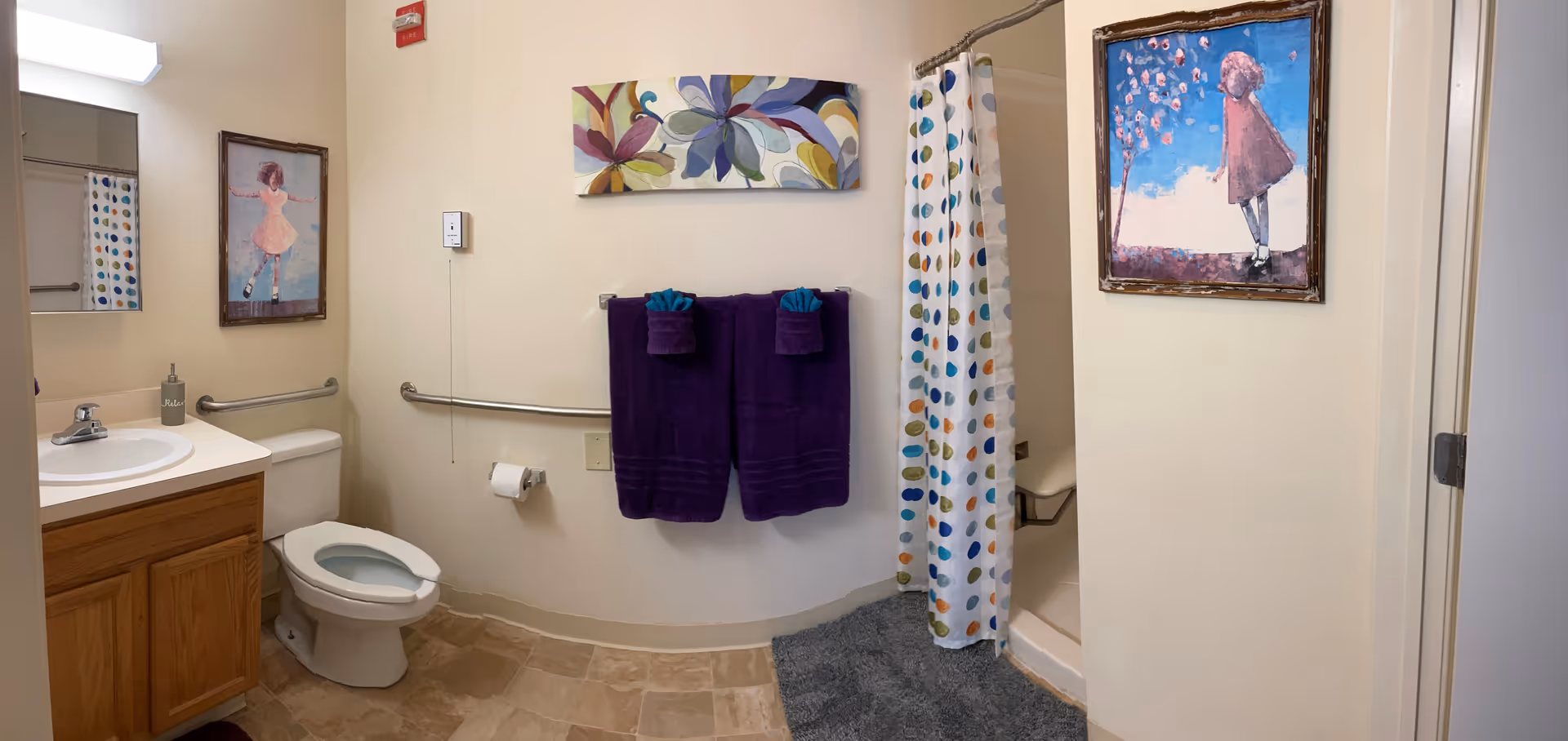 A bathroom with a toilet, sink with wooden cabinet, and a shower with a colorful polka dot curtain. Two purple towels are hanging on a towel rack on the wall. The walls are decorated with three framed artworks featuring children. The floor has beige tiles and there is a gray bath mat in front of the shower.