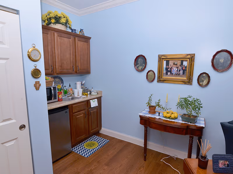 A small kitchenette area with light blue walls, wooden cabinets, a countertop with various items including a microwave, cups, and a basket. There is a small refrigerator under the counter. On the right side, a wooden table with potted plants, lemons, and candles sits against the wall decorated with framed pictures and portraits. The floor is wooden, and there are decorative items on the wall and a mat on the floor.