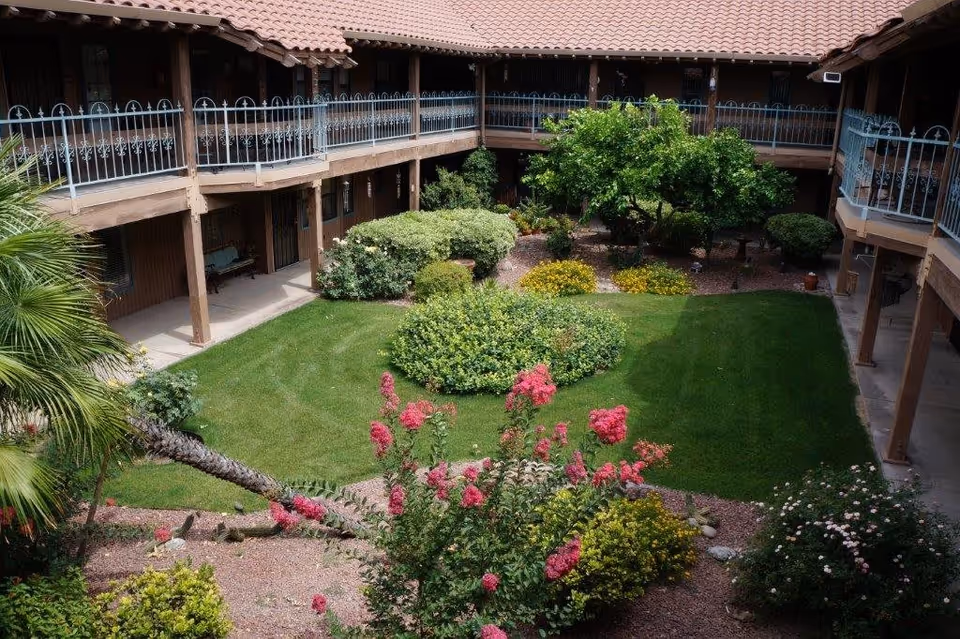 View of a landscaped courtyard garden with green grass, various bushes, flowering plants, and a tree, surrounded by a two-story building with a tiled roof and balconies with metal railings.
