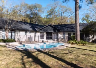 Outdoor view of a single-story building with a black roof and white walls, featuring a fenced-in swimming pool in the foreground surrounded by grass and trees.
