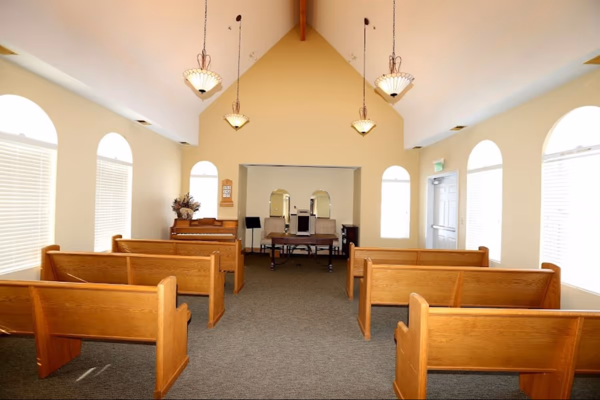 Interior view of a small chapel or meeting room with wooden pews arranged in rows facing a small stage area with two chairs, a lectern, and a piano. The room has high vaulted ceilings with hanging pendant lights and arched windows with blinds letting in natural light.