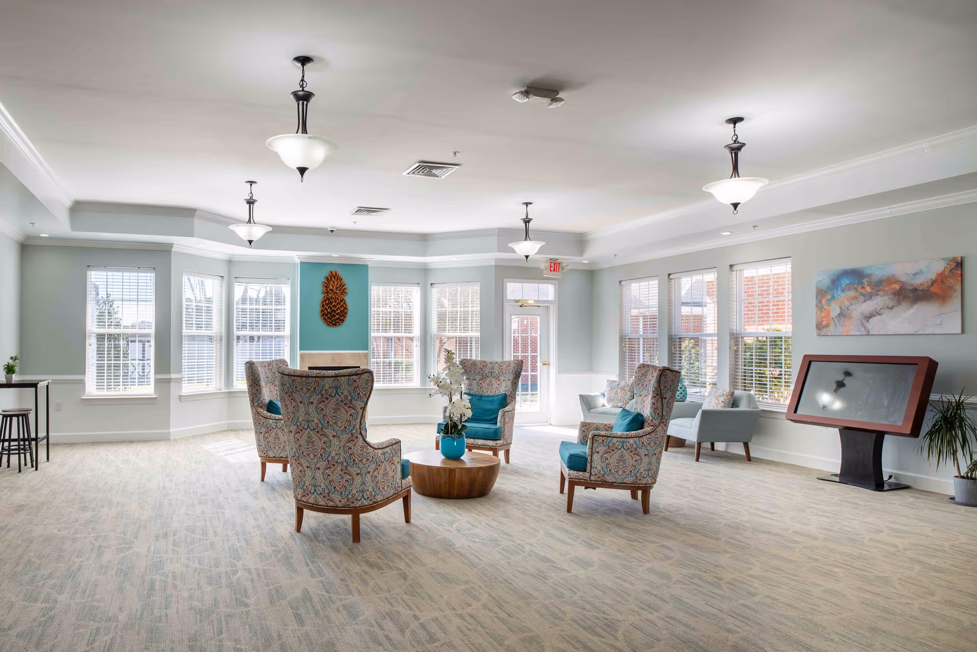 Bright senior living community lounge with patterned armchairs arranged around a low wooden table, large windows, and a touchscreen kiosk.