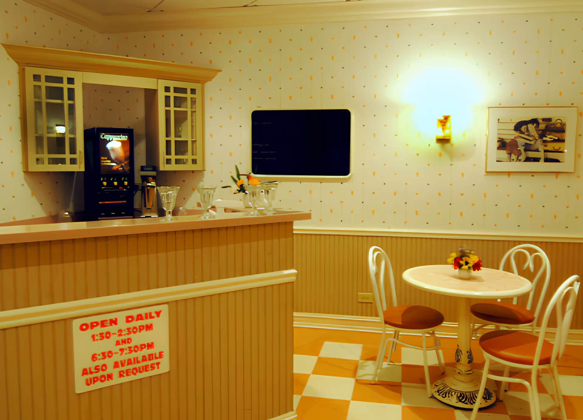 A small dining area with a round table and four chairs with orange cushions. There is a counter with a coffee machine and glassware on it. The walls have light-colored wallpaper with small orange patterns, a wall-mounted light fixture, and a framed picture. A sign on the counter indicates daily open hours.